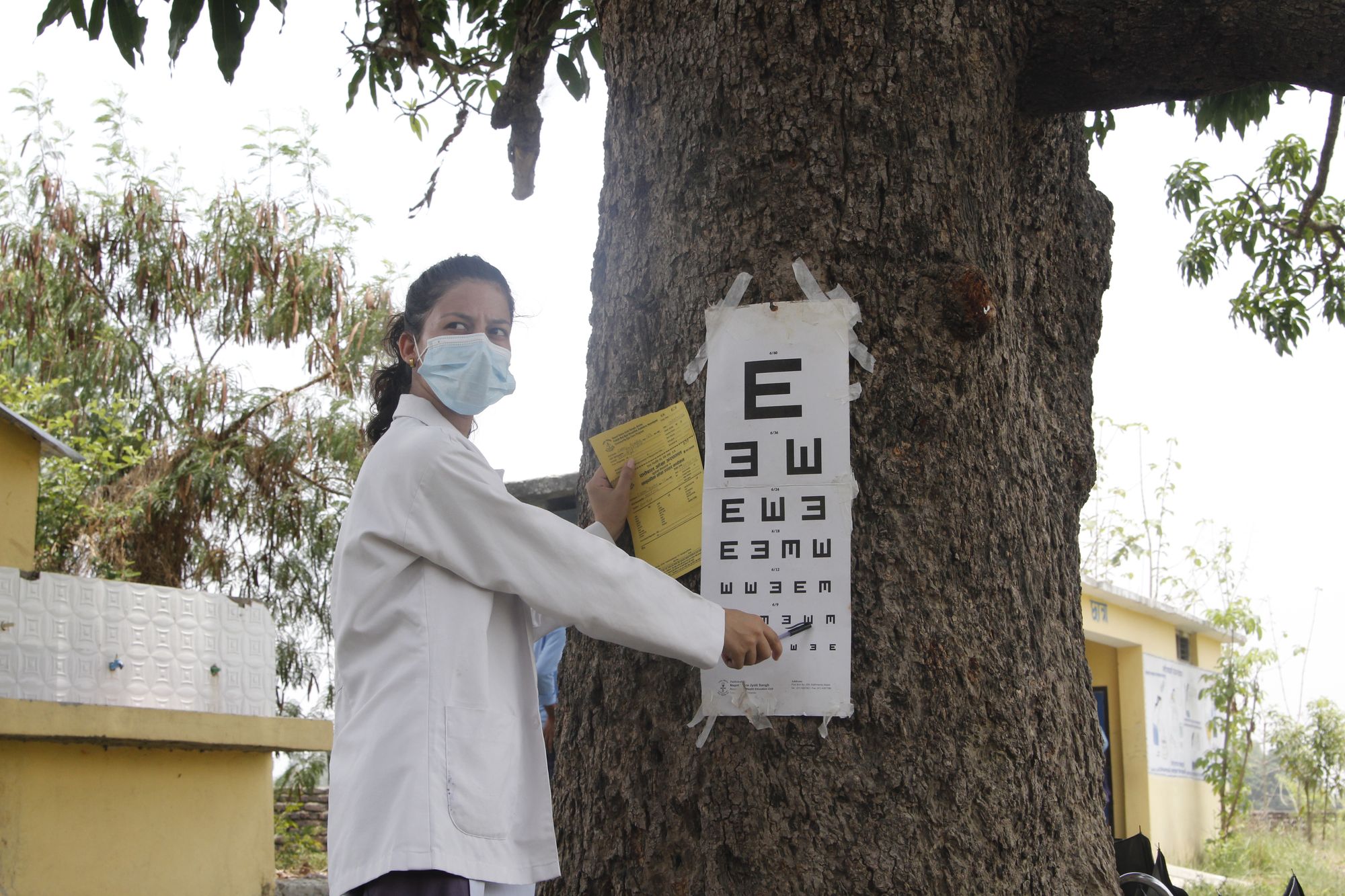 A female trainee doctor in a white lab coat pointing at a chart to conduct an eye test at a Tej Kohli & Ruit outreach camp
